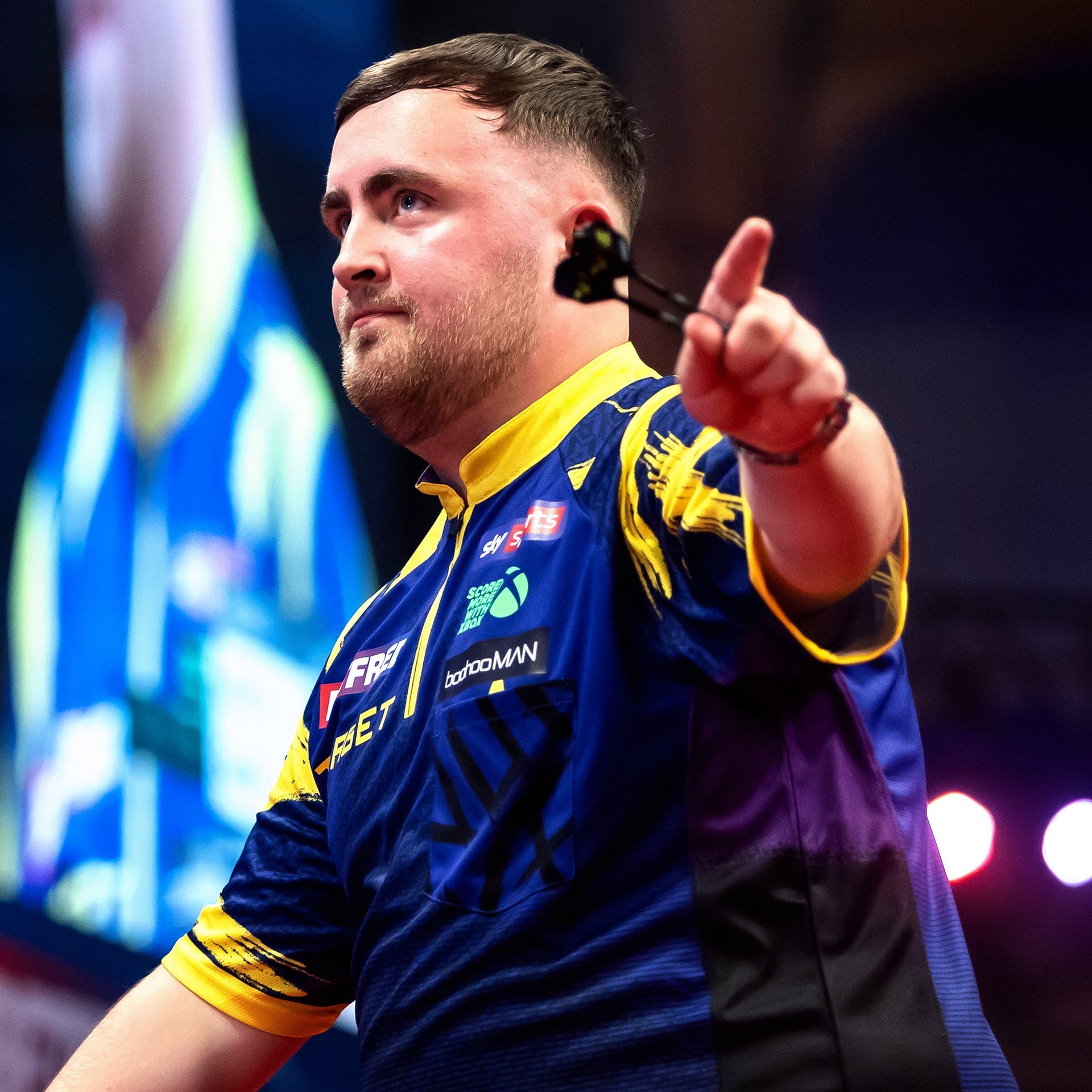 Luke Littler in a purple and yellow sports jersey pointing forward with a blurred background