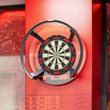 A wall-mounted dartboard with a clear protective circular cover and black mounting hardware, set against a red-orange illuminated wall with the word TARGET visible.