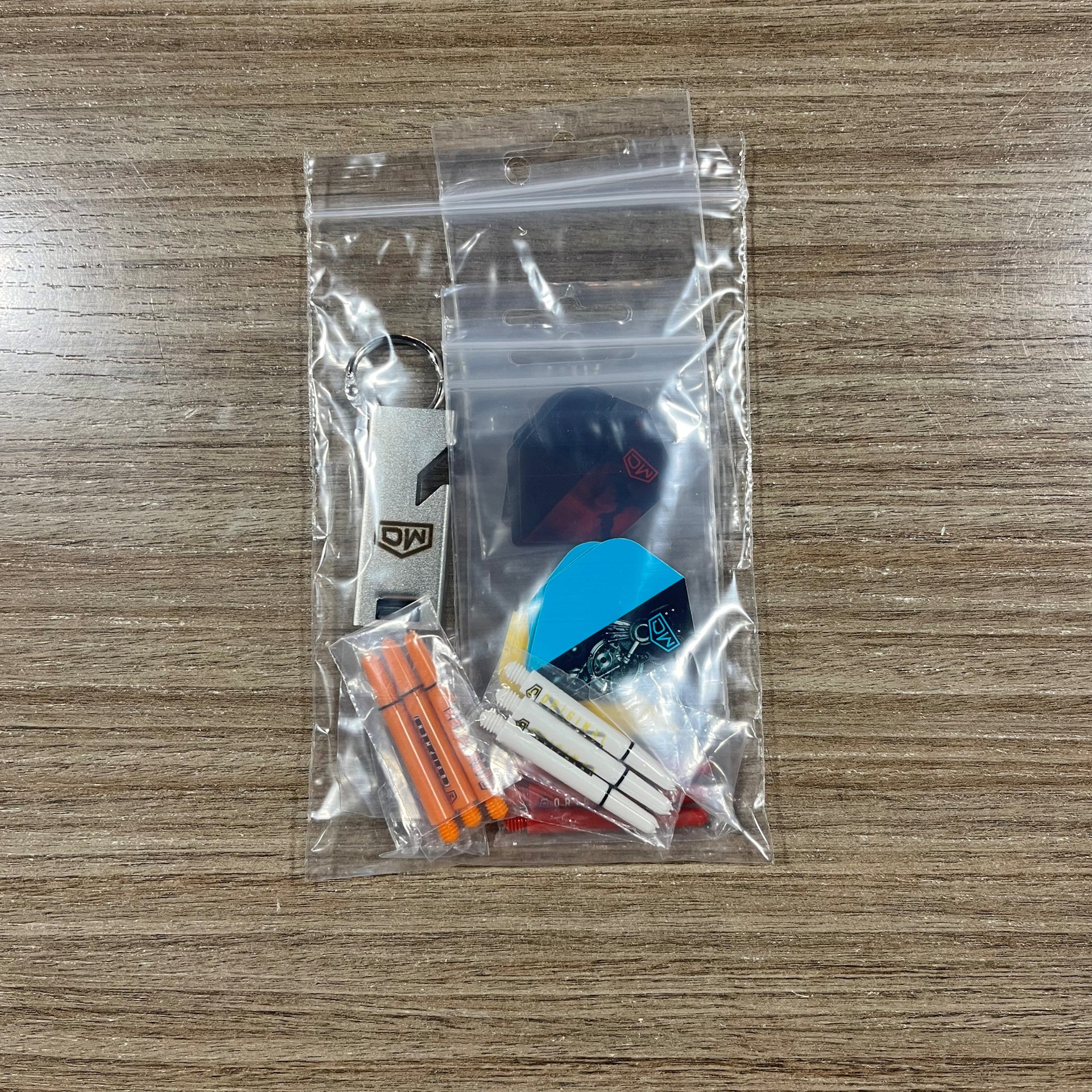 A clear plastic zip-top bag containing a small dart keychain set on a wood-grain tabletop. The set includes assorted dart Accessory Pack. Side B